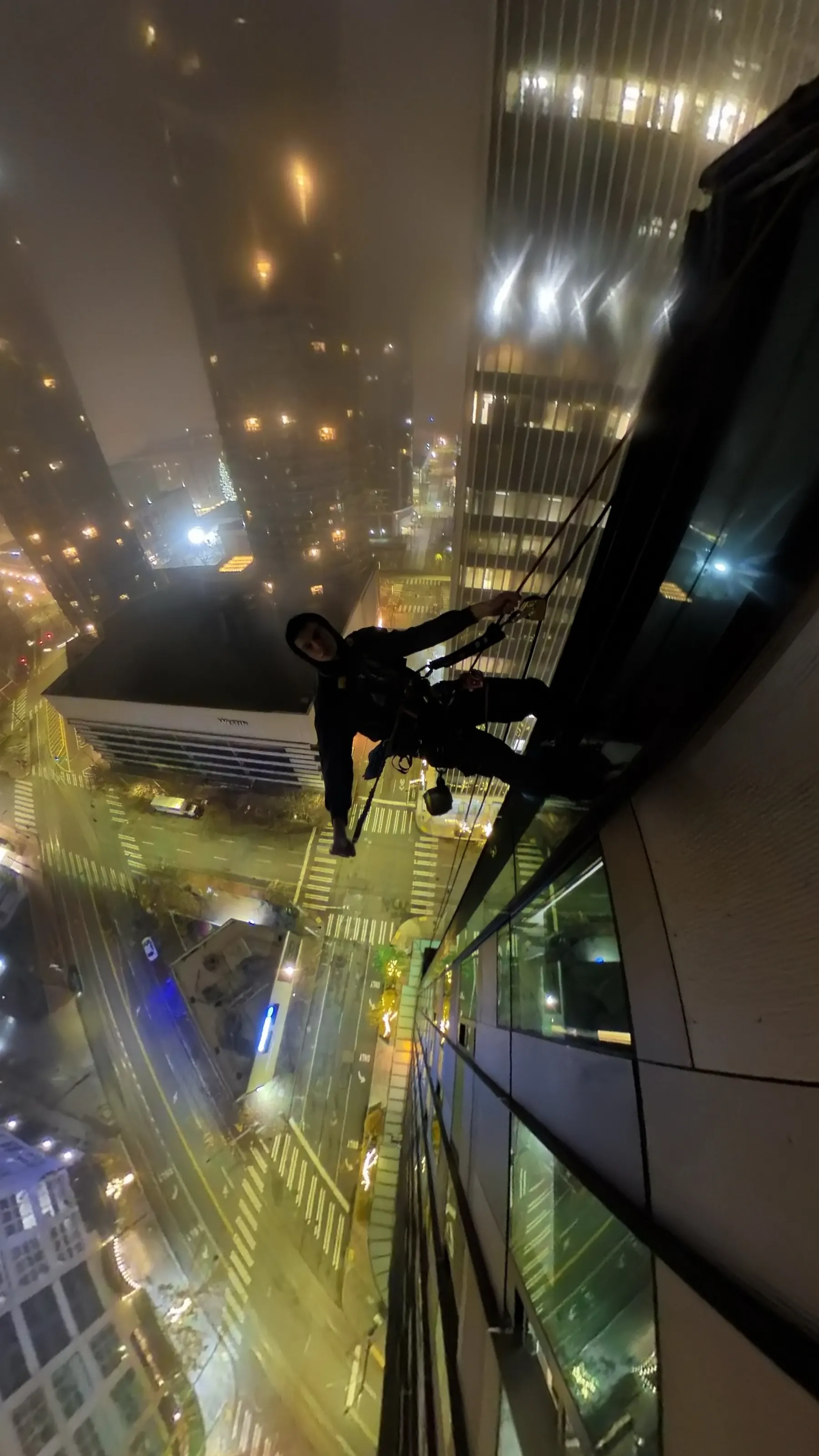 Technician on ropes at night beside a lit high-rise, city streets below